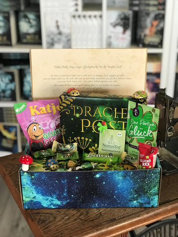 Das Produktbild zeigt vorn eine geöffnete Drachenpost in blauem Schuppen-Design. Man sieht den in den Boxdeckel geklebten Brief zum Thema Glück und in der Box ein in ein grünes Drachenpost-Poster verpacktes Buch und zum Thema passende Goodies - Süßigkeiten und andere Kleinigkeiten. Im Hintergrund sieht man die Bücherregale des Verlagsladens.